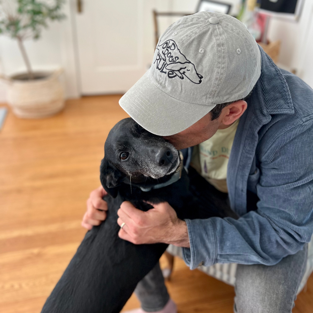 Man wearing the beige Old Dogs Rule embroidered cap hugging a senior black dog
