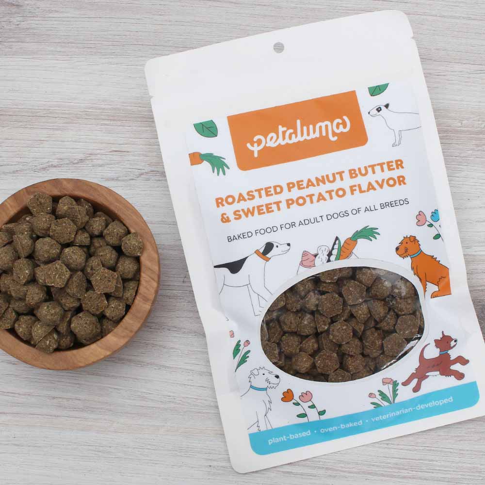 A package of Petaluma brand dog treats with roasted peanut butter and sweet potato flavor. The package is open, showing the treats inside, next to a wooden bowl filled with the treats.